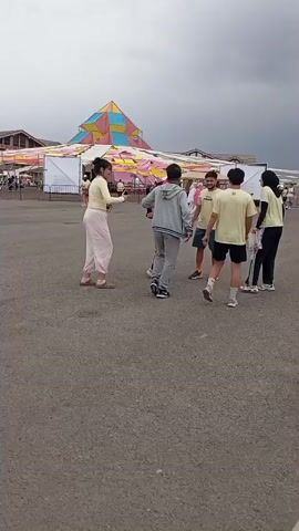 A group of six young people are standing in a circle on an asphalt surface. In the background, there is a large, colorful tent-like structure, as well as some smaller tents and structures. The sky is cloudy. The group appears to be interacting, possibly playing a game or engaged in conversation. One person, wearing a grey hoodie and sweatpants, is in the center of the circle, facing towards the other individuals. The other five people are standing around them, some of them facing inwards. Their clothing is casual, consisting of t-shirts, shorts, and pants. Some people are holding objects, possibly phones or papers.