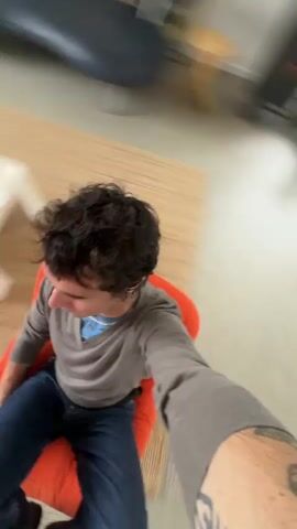 Here's a concise description of the video:

A man with curly dark hair sits in an orange chair. He wears a grey long-sleeved shirt, a blue shirt underneath, and jeans. He's holding the camera at arm's length, giving a first-person perspective of himself sitting.  The background shows a living room with a rug, furniture, and white walls. The camera moves slightly, showing more of the room.  There appears to be music playing in the background.