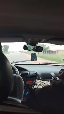 The video shows the interior of a car from a rear passenger's perspective, looking forward. The driver, a person with dark skin and hair, is visible in the driver's seat, wearing a grey shirt. A mobile phone is mounted on the dashboard in front of the windshield. Another person is in the front passenger seat, holding a mobile phone. The car is driving on a paved road with houses and vegetation visible on either side. The sky is overcast. The time stamp indicates the video is from 00:00 to 00:05.
