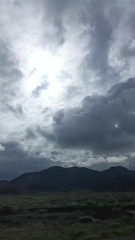 The video shows a landscape view with mountains in the background and a field of grass in the foreground. The sky is filled with dramatic clouds and the sun is visible through a break in the clouds. The overall atmosphere is moody and overcast.