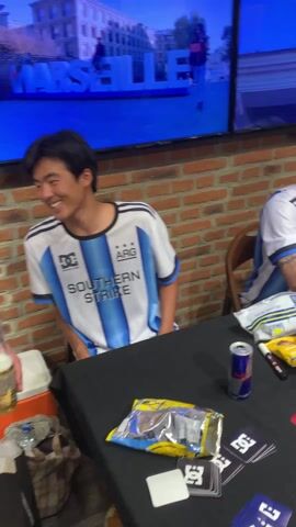 The video shows a group of people gathered around a table in what appears to be a retail setting, possibly a shoe store. Three men are wearing matching white and blue soccer jerseys with the word "SOUTHERN" on them, sitting at a table with snacks, drinks, and DC-branded items like stickers and cards. Two people are taking photos of the men. One photographer is using a large camera with a flash, while the other is using a mobile phone. Shelves filled with shoes are visible in the background. A television screen displays a view of an outdoor plaza. The atmosphere seems casual and friendly, suggesting an autograph session or meet-and-greet event.