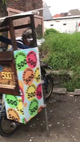 The video shows a street vendor's cart selling what appears to be a type of meatball called "PENTOL." The cart is colorful with a rainbow umbrella providing shade. A person, likely the vendor, is seated behind the cart, wearing a dark shirt and a cap. The cart has wheels and is situated on an outdoor paved surface, possibly a street or sidewalk. There is a wall made of bricks in the background, overgrown with green grass and vegetation. The scene suggests a typical outdoor market or roadside food stall.