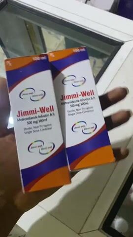 The video shows a person's hands holding two identical boxes of Jimmi-Well Metronidazole Infusion B.P. 500 mg/100ml. The boxes are orange and blue and have the product name and details printed on them. The background appears to be an indoor environment with a light-colored surface. The lighting is dim, and the focus is on the boxes.