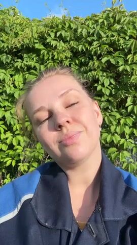 A young woman with blonde hair pulled back is looking at the camera. She is wearing a dark blue jacket with a light blue stripe. She is standing in front of a green leafy hedge with a blue sky above. The woman blinks a few times and smiles.