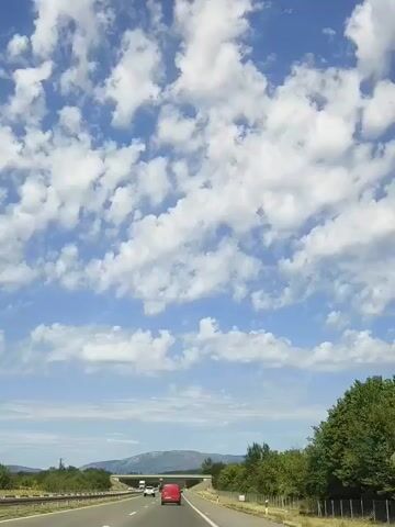 The video shows a view from the perspective of a vehicle driving on a highway. The sky is a bright blue with numerous white, fluffy cumulus clouds scattered across it. The highway itself has at least two lanes of traffic, with other vehicles visible ahead, including a red car in the foreground and a white truck further ahead. To the right of the highway, there is a dense line of green trees and vegetation, with a fence running alongside the road. In the distance, there are hills or mountains visible under the horizon line. The overall impression is a sunny day while traveling on a road.