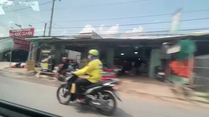 The video, taken from inside a moving vehicle, shows a street scene in Vietnam. The camera faces toward the right side of the street, capturing various buildings and businesses. Some buildings are open-air workshops or garages, while others appear to be residences. One sign reads "CƠ SỞ NGỌC QUÝ PHÁT." A person wearing a conical hat is riding a bicycle on the street. Another person on a scooter passes by. Power lines and poles run along the street. The sky is blue with some clouds.