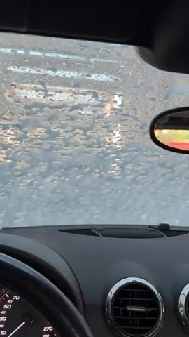 The video is shot from inside a car, looking out through the windshield. The windshield is covered in soap suds and water, suggesting the car is going through a car wash. We can see a portion of the car's dashboard, including the steering wheel and an air vent. A side mirror is also visible. The lighting outside the car suggests it is daytime, possibly late afternoon, with sunlight reflecting off the wet windshield.