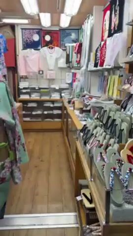 The video shows the inside of a shop selling traditional Japanese items. The shop is small, with a narrow aisle running through it. On either side of the aisle are displays of kimonos, fans, paintings, and other items. A price tag on a mint green kimono shows a price of 7700 Yen. The camera pans to show a customer browsing through the merchandise. Then, the camera moves to a view of a street lined with shops. The street is crowded with people, and many red lanterns and floral decorations are hung overhead. The time is night.