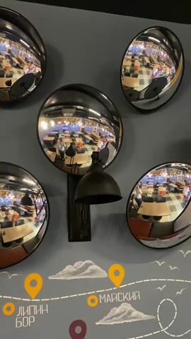 The video shows a wall decorated with several convex mirrors and a black industrial-style lamp. The mirrors reflect a brightly lit room, possibly a restaurant or cafe, with tables and chairs. People can be seen reflected in the mirrors, some standing and some sitting. Below the mirrors, the wall is painted with a map-like design featuring clouds, location markers, and text in Russian, including "ЛИПИН БОР", "МЕДВЕЖЬЕГОРСК", and "МАЙСКИЙ". The video pans slightly to the right, giving a closer view of the lamp and the arrangement of the mirrors.