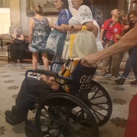 The video depicts an indoor scene inside what appears to be a church or cathedral, as suggested by the elaborate architecture, interior design, and the presence of religious figures and artifacts.

In the foreground, a person in a wheelchair is being pushed through the crowd. The wheelchair has the letters "GAL" written on the back.

As the camera moves, it focuses on a woman sitting in a confessional booth, facing a priest who is blessing her.

The surrounding area is filled with other people, some of whom are walking around, while others are standing or sitting. A woman is carrying a yellow bag. The floor has a patterned tile design.