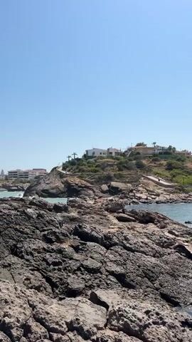 The video shows a rocky shoreline with a clear blue sky. In the background, there are white buildings on a hill overlooking the sea. The water appears calm and the sun is shining. There are no people visible in the video. The focus is on the natural landscape with the man-made structures in the distance. The camera is positioned low, emphasizing the texture and ruggedness of the rocks in the foreground. The overall impression is one of a serene coastal environment on a bright, sunny day.