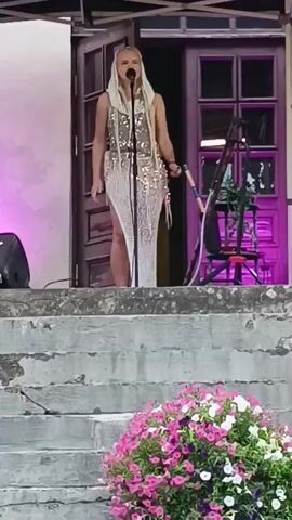 A woman with blonde hair in a white dress with sparkly accents stands on a stage and sings. She is holding a stick. There are stairs in front of her with pink and white flowers at the bottom. There is a doorway behind her with purple lighting.