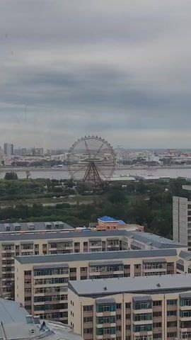 The video shows a cityscape under a cloudy sky. In the distance, a large Ferris wheel is visible, with a river and greenery separating it from the foreground. The foreground is dominated by several apartment buildings of varying heights and architectural styles. Some buildings are light-colored with many windows and balconies, while others have more decorative elements like patterned facades and peaked roofs. The overall impression is of a densely populated urban area with a mix of modern and older residential architecture. There are no people or specific actions clearly depicted in the video.