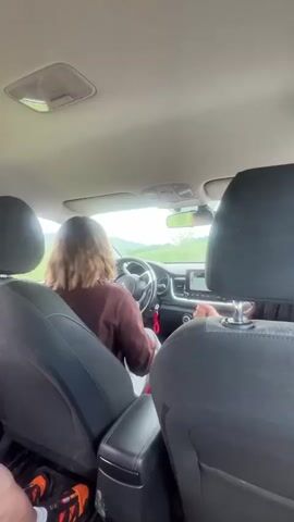 The interior of a car driving on a scenic road is shown in this video. A woman with shoulder-length brown hair and sunglasses is driving and singing along to music. She is wearing a brown shirt. A passenger in the back of the car is visible, with long dark hair. The passenger is filming a video of themself while lip-syncing to the music and holding a pink phone. The landscape outside the windows includes green hills and a cloudy sky.