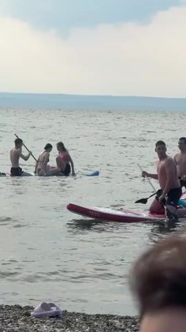 Four individuals are paddleboarding on a body of water. Two individuals are on one paddleboard, and two individuals are on a separate paddleboard. One of the individuals on the second paddleboard is paddling with a paddle. The water is calm with small waves. The sky is cloudy. In the foreground, there is a beach with pebbles and a bird.