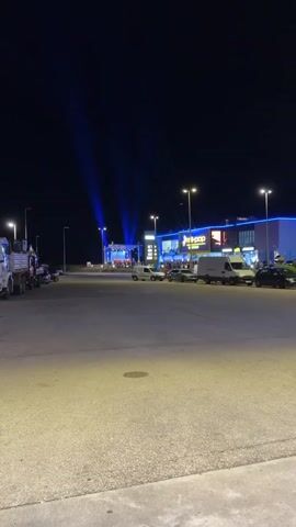 The video shows an outdoor stage set up in front of a building, possibly a shopping center, illuminated with blue lights and spotlights. There appears to be a choir or musical group performing on the stage, observed by an audience gathered around the stage area. A large electronic sign displays gas prices next to the stage. Several cars and vans are parked nearby, suggesting a gathering or event is taking place. The scene is set at night.