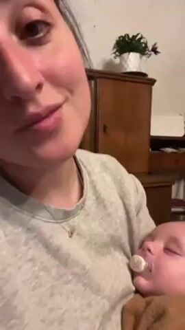 A person holds a sleeping baby, who has a pacifier in their mouth. The person holding the baby has a gray shirt and a necklace. The baby is wrapped in a brown blanket. Behind the person and baby is a wooden cabinet with a plant on top of it. There is a white object on the cabinet, as well.
