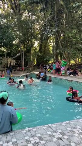 This video depicts an outdoor swimming pool area surrounded by lush trees and vegetation. The pool is filled with clear blue water and several people, predominantly children, are engaged in various water activities. Some children are swimming, while others are playing with inflatable rings. Two children are prominently featured near the foreground, one with a green inflatable ring and another with a black inflatable ring. In the background, a group of adults are seated on the poolside or standing, observing the children. There is also some inflatable play equipment visible on the side of the pool, and several trees provide a canopy of shade over the area. The overall atmosphere appears to be one of leisure and recreation.