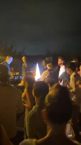 A group of people are gathered outdoors at night. A person in a yellow cap and dark jacket is standing and appears to be speaking into a microphone, holding a reflective object in their left hand. Several people are facing the person speaking, and others are looking in different directions. There is a bright light source in the center of the image. The background is dark, suggesting it is nighttime. The environment appears to be a backyard or outdoor gathering space.