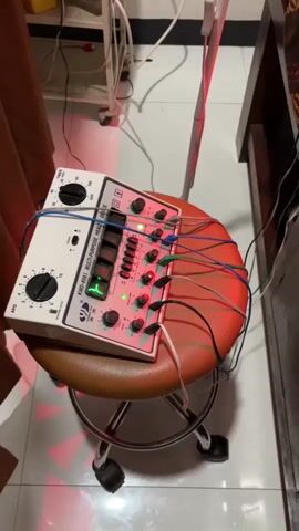 The video shows a white medical device with numerous wires connected to it. The device is labeled "KWD-801 MULTI-PURPOSE HEALTH". It is placed on a brown circular stool with wheels. Several small lights on the device are illuminated. The background is blurred, but appears to be an indoor setting with some medical equipment and furniture. Red light is also visible on the stool and surrounding area, possibly from a therapeutic lamp.