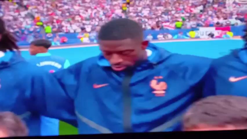 The video shows a close-up of two men in blue jackets with an orange crest. They are standing side-by-side, with their arms around each other's shoulders. They are looking forward, and their faces are visible. The man on the right has a serious expression, and the man on the left is looking to the side. In the background, there is a crowd of people in the stands, and a blue field. The video is shaky, and the lighting is dim. The overall impression is of a moment of reflection or solidarity between the two men.