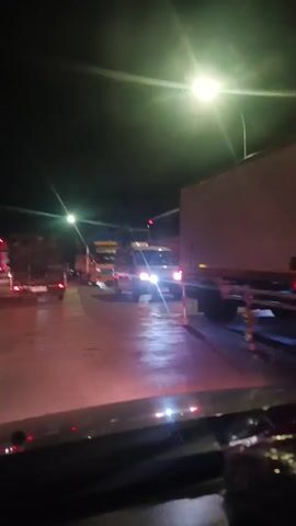 The video shows a traffic scene at night. There are several trucks and a van on a wet road. Streetlights illuminate the scene. The camera is inside a vehicle, looking forward. The lighting is dim, and there are lens flares from the headlights.