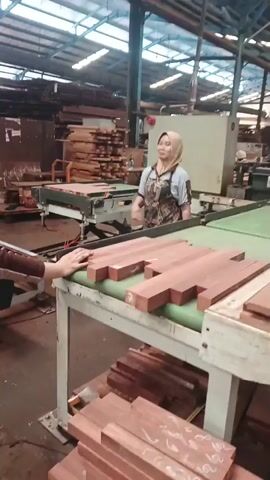 Two women, both wearing headscarves and aprons, are in a woodworking factory. The woman on the right is operating a machine, pressing buttons. The woman on the left is standing next to her, with her hands resting on a conveyor belt with wooden planks. There are stacks of wood in the background and foreground. The environment appears to be a workshop or factory floor.
