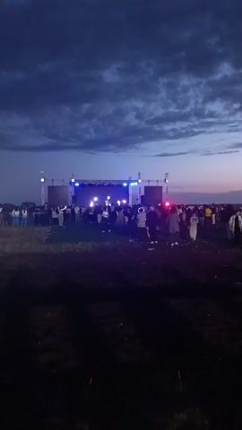 The video shows a large crowd of people gathered in an open field at dusk, facing a brightly lit stage. The stage is set up with a large screen and several speakers, suggesting a musical performance or event is taking place. Colored lights, predominantly blue and pink, illuminate the stage and the surrounding area, casting a vibrant glow against the darkening sky.

The sky is filled with dramatic, cloudy formations, indicating either sunset or a partly cloudy evening. The overall atmosphere appears energetic and festive, with people milling about and enjoying the event. The foreground of the video is dark, suggesting the camera is positioned at ground level in an unlit area of the field, possibly on a dirt track or uneven terrain, with tire tracks visible.

The subjects are a diverse group of people of various ages, dressed in casual clothing. They are interacting with each other and engaging with the spectacle on stage. There are no visible signs or text within the frame. The setting is a rural or semi-rural outdoor location, likely in Kazakhstan given the location context, with a vast open space. The time of day is clearly evening, transitioning from twilight into night.