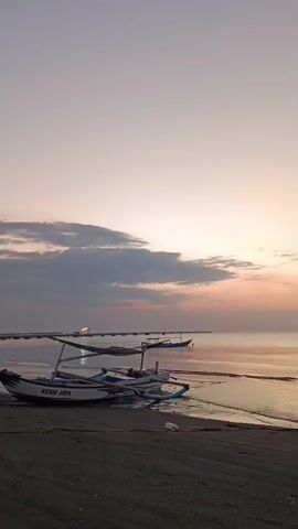 The video shows a serene beach scene at dusk. In the foreground, a few traditional fishing boats are pulled up onto the dark sand. The sea is calm with gentle waves lapping at the shore. In the distance, a pier stretches out into the water, with a few lights visible. The sky is a soft gradient of pale orange and pink, with scattered clouds. The overall mood is peaceful and tranquil.