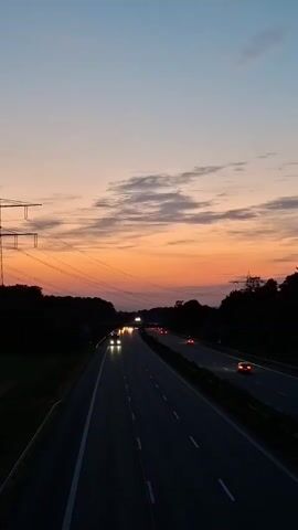 The video shows a highway at dusk. The sky is a gradient of blue and orange with scattered clouds. The highway is dark with white lane dividers, and there are cars with their headlights on driving in both directions. On the left side of the highway, there are tall power lines and a large metal pylon. Trees line the sides of the highway, appearing dark silhouettes against the colorful sky. The overall impression is one of travel and the transition from day to night.