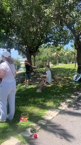 The video shows a group of people in a park on a sunny day, participating in various outdoor activities.  There are several adults and children present.  Some are playing ring toss, while others are engaged in activities on a makeshift obstacle course marked by flags and tape. A father is helping his son navigate the course. In the background, there is a lake with the skyline of a city visible on the opposite shore.  The park has grassy areas, trees, and paved paths.  One woman is talking on her phone.