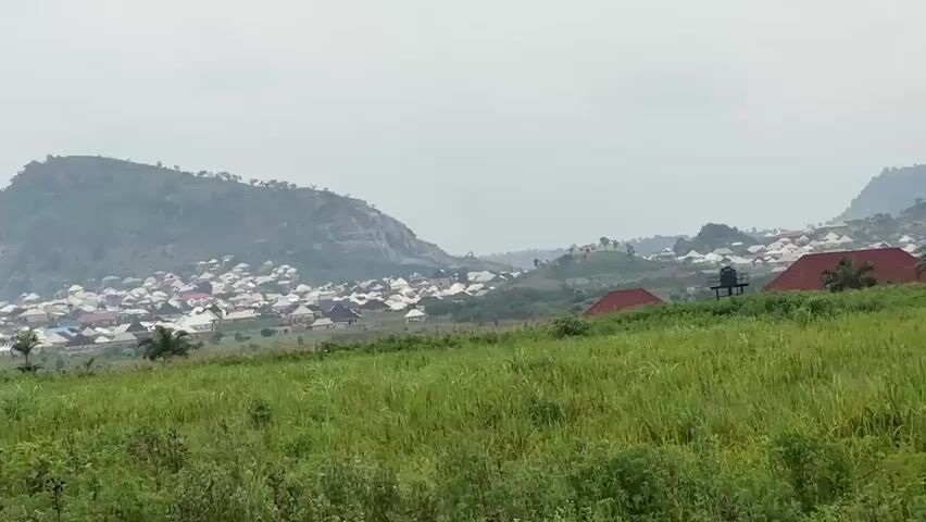This is a wide-angle video of a rural landscape on an overcast day. In the foreground, there is a field of tall, green grass with some lower-lying green plants at the very bottom of the frame. In the middle ground, there is a small collection of buildings, some with red roofs. One building has a water tank on top. There are also several palm trees scattered throughout this area. In the background, there is a town or village spread out across rolling hills, with mountains rising behind it. Some of the buildings in the town are white, and some have darker roofs. There is a large, long building with a reddish-brown roof situated in the middle background. The sky is cloudy and grey. The camera pans slowly from left to right.