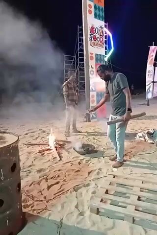 At night, two men are on a sandy surface. In the foreground, a man in a dark t-shirt and light pants initially holds a wooden stick near a small fire producing smoke on the sand. He then places a small clear bottle containing yellow liquid onto a dark metal barrel with circular holes, which is positioned to his left. He then turns and walks towards the right, away from the fire. Another man in a plaid shirt and light pants stands behind the fire, partially obscured by smoke. In the background, a tall, illuminated sign reads "GOA FOOD FESTIVAL" with colorful patterns. A wooden pallet lies on the sand to the right.