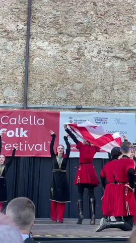 A group of dancers are performing on a stage in front of a stone wall. The dancers are wearing traditional folk costumes. Some are dressed in black and gold outfits with red leggings, while others are in vibrant red and white attire, one person prominently displaying a Georgian flag. The stage is set up outdoors, with a banner behind the performers that reads "Calella Folk Festival." Additional text on the banner includes "Ajuntament de Calella," "Fortuni Viateges," and "Folklore Festival Association" with a social media handle and website. The overall scene suggests an outdoor folk festival taking place during the daytime. The dancers appear energetic and engaged in their performance, with some smiling and raising their arms. The setting appears to be a public space, possibly a town square or park in Calella, Spain, given the visible text.