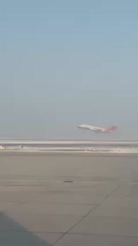 The video shows an airplane taking off from a runway. The plane is white with red accents. The runway appears to be mostly grey concrete with snow on either side. The sky is a pale blue. The video is shot from ground level.