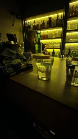 The video shows a bar counter with two shot glasses with a lemon slice on the rim. A phone lies next to them. Two bartenders work behind the counter, which is lined with various liquor bottles backlit by warm yellow lights. A woman with long nails squeezes a lemon slice over one of the shot glasses. Another person's hand briefly reaches toward the shots.