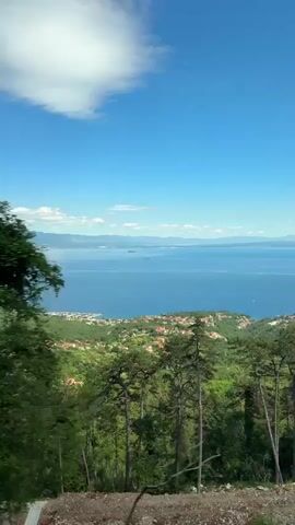 The video shows a wide shot of a coastline with a clear blue sky and some clouds. In the foreground, there are trees and green foliage. In the middle ground, there is a coastal town with red-tiled roofs nestled on a hill overlooking a large body of blue water. In the distance, there are mountains and a ship on the water. The scene appears to be captured from a moving vehicle, as evidenced by the slight blur of the foreground foliage.