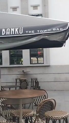 The video pans across an outdoor seating area of a establishment. In the foreground, there are several tables and chairs, some of which are bar stools with woven seats. The tables are round and appear to be made of metal. Above the seating area, a large black umbrella with white lettering that reads "BANKU THE TOWN BAR" is visible. In the background, there is a building with large windows and a doorway. A large potted plant with broad leaves stands near the entrance. The overall environment suggests a commercial establishment, possibly a restaurant or bar, with outdoor seating.