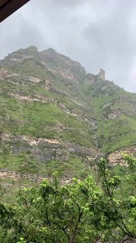 The video shows a cloudy, overcast day with a view of a lush green mountain range. The mountain slopes are layered with rock formations and covered in vegetation. In the foreground, there are trees with green leaves. The overall impression is a natural, rugged landscape under a gray sky, suggesting a potentially damp or rainy environment. There are no people or distinct man-made objects visible apart from possibly a railing at the very bottom edge of the frame. The camera appears to be stationary.
