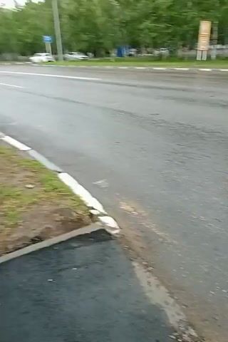 This media file depicts a wet asphalt road, likely after rain. The scene appears to be a street or intersection with some sparse vegetation and trees in the background. There are no discernible people or animals visible.

Objects in the scene include a yellow vehicle, possibly a van or bus, and some traffic signals. The road surface is reflective, indicating the presence of water. The overall lighting suggests it is daytime, but the sky is overcast, contributing to a somewhat muted color palette.

The primary activity is the visual recording of a street scene during or immediately after rainfall. The motion of the camera is generally horizontal, suggesting it might be mounted on a moving vehicle or held by someone walking.

Notable details include the wetness of the road, the overcast sky, and the presence of traffic infrastructure. The location is identified as Kostanay, Kazakhstan. There is no visible text within the frame of the video. The mood of the video is neutral, focusing on the environmental conditions and the urban landscape.