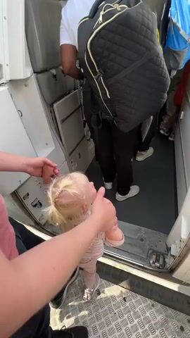 The video shows the interior of an airplane aisle. A young child with blonde hair is being held by a person whose arm is visible in the foreground. The child is looking up towards other passengers in the aisle. Behind the child, several people are standing, some with backpacks. One person is wearing a white t-shirt and a black backpack. Another person has a large blue backpack. The aisle appears to be narrow, and the passengers seem to be boarding or disembarking. The airplane cabin has overhead lights and luggage compartments. The windows on the right side of the plane show an "ISRAIR" logo. The floor of the aisle is dark gray.