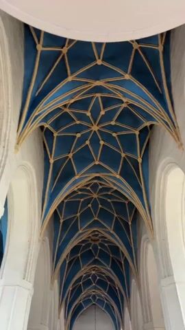 The video pans upwards to reveal a highly ornate ceiling of a building, likely a church or cathedral. The ceiling is a vaulted structure with intricate ribbed vaulting. The vaulting bays are a deep blue color, divided by thin, golden geometric lines that form star-like patterns and diamond shapes. The ribs themselves are also gold and appear to be raised or slightly three-dimensional. The white walls of the structure are visible on either side, with large archways that are part of the architectural design. The perspective of the camera suggests it is positioned in the center of a long hall or nave, looking up towards the apex of the ceiling. The lighting is even, suggesting an interior space, and there are no visible figures or active events occurring within the frame. The overall impression is one of architectural grandeur and detailed craftsmanship.