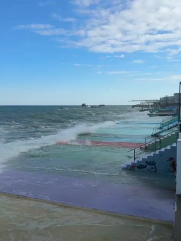 The video shows a rocky coastline with waves crashing against the shore. There is a colorful swimming pool area with steps leading down to it. The sky is blue with some clouds, and the water is choppy. The scene appears to be in a place where the sea meets land, and the pool is designed to be filled by the ocean.