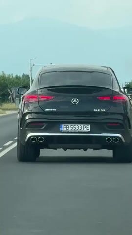 This video shows the rear view of a black Mercedes-Benz GLE 53 AMG Coupe driving on a road. The car has a Bulgarian license plate that reads "PB 5533 PE". In the background, there are trees and a signpost with Bulgarian text and numbers indicating directions to "Царимир" (Tsarimir) and "Неделево" (Nedelevo), with distances of 5 and 13 respectively. The sky is overcast, suggesting a cloudy day. The setting appears to be a rural or suburban area in Bulgaria, possibly near Graf Ignatievo, as indicated. There are no people visible in the video, and the focus is solely on the car and its surroundings. The time of day appears to be daytime due to the ambient light. The overall tone is neutral, simply documenting the car's movement on the road.