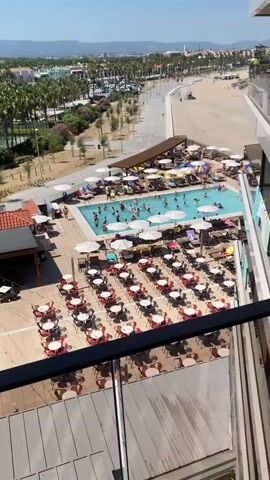 This is a high-angle aerial video of a resort pool area on a sunny day. In the foreground, there are rows of red and white tables with chairs set up on a tiled patio. Beyond the patio is a rectangular swimming pool filled with many people swimming, playing, and wading. There are numerous lounge chairs and umbrellas surrounding the pool, some occupied by sunbathers. A man is seen walking along the edge of the pool. In the background, there is a beach with sand and waves, lined by palm trees and a street with cars. Further in the distance, there are buildings and a hazy landscape. The overall scene depicts a lively resort atmosphere with recreational activities in full swing.
