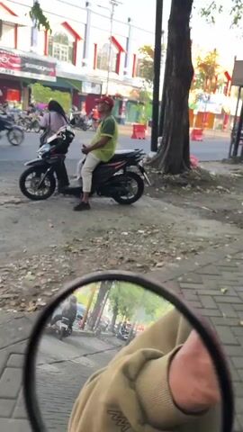 A man in a green shirt and white pants sits on a black motorcycle. He is wearing a red cap. Another person, wearing dark clothing and a pink headscarf, stands next to the motorcycle. In the background, a yellow vehicle, possibly a van, is visible. The scene appears to be outdoors on a street with buildings in the distance. The lighting suggests it is daytime. The camera is positioned low, possibly from the perspective of someone on another motorcycle or vehicle.
