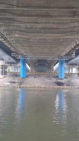 The camera is stationary and positioned under a large concrete bridge. The view is across a body of water, which reflects the blue supports of the bridge. The water appears calm. A red barge slowly moves from right to left, partly obscuring the view of the bridge's supporting structure. The barge's hull is visible, with some equipment on the deck. The area under the bridge is mostly gray concrete with blue-painted pillars and a black and yellow caution stripe along a low wall.