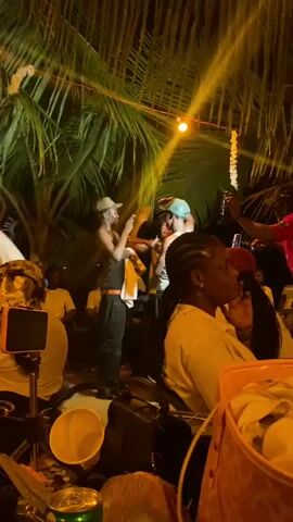 The video shows a group of people outdoors at night, possibly at a party or event. Two shirtless men are dancing near a palm tree adorned with string lights and flower garlands. Other people are seated and standing around the dance area, some watching, some recording on their phones. The light is warm and seems to come from overhead spotlights. There are also various objects scattered around, including a bucket, a can, and a coconut. The general impression is of a festive and lively atmosphere.