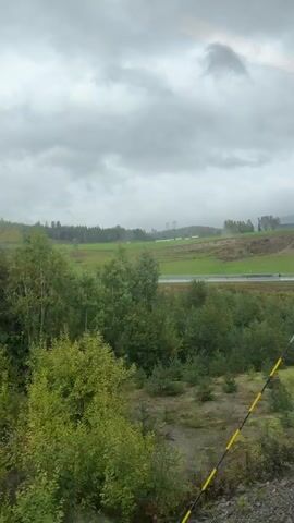The video shows a landscape view from a moving vehicle, likely a train, on a cloudy day. In the foreground, there are trees and bushes. A road with guardrails runs horizontally across the middle ground. Beyond the road, there is a green field and a wooded area with trees on a slope. In the distance, there are hills and power lines. The overall atmosphere is somewhat overcast and natural.