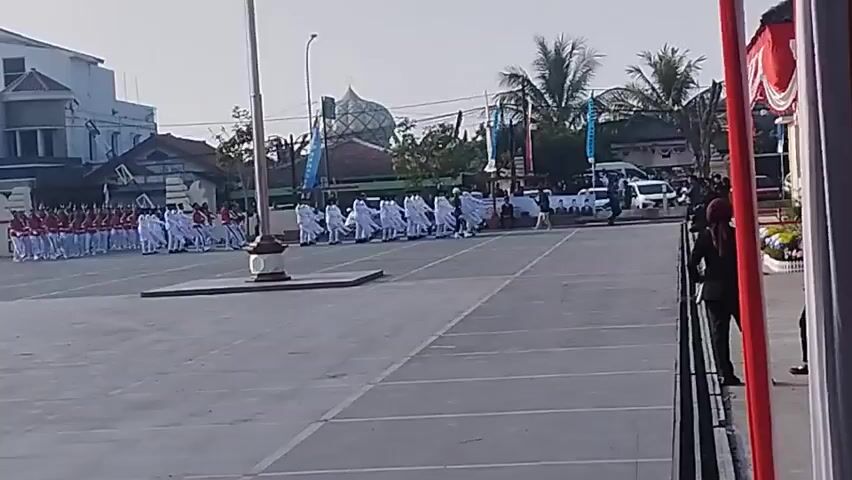 The video shows a large, paved outdoor area, likely a plaza or parade ground, with a flagpole in the foreground. On the left side of the frame, a group of individuals in red and white uniforms are standing in formation. In the middle ground, a group of people in white robes are marching in a line, some carrying flags or banners. In the background, buildings, palm trees, and vehicles are visible. The sky is clear and the lighting suggests it is daytime, possibly morning or late afternoon. The overall impression is of an organized event or ceremony.